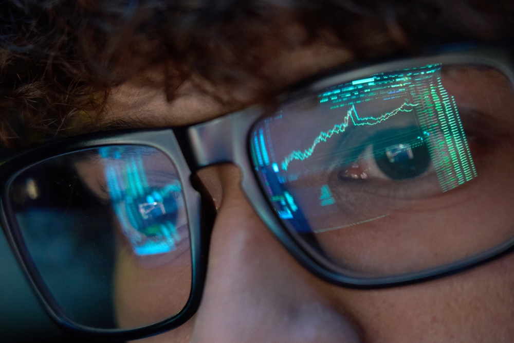 trader wearing glasses looking at computer screen with trading charts reflecting in eyeglasses