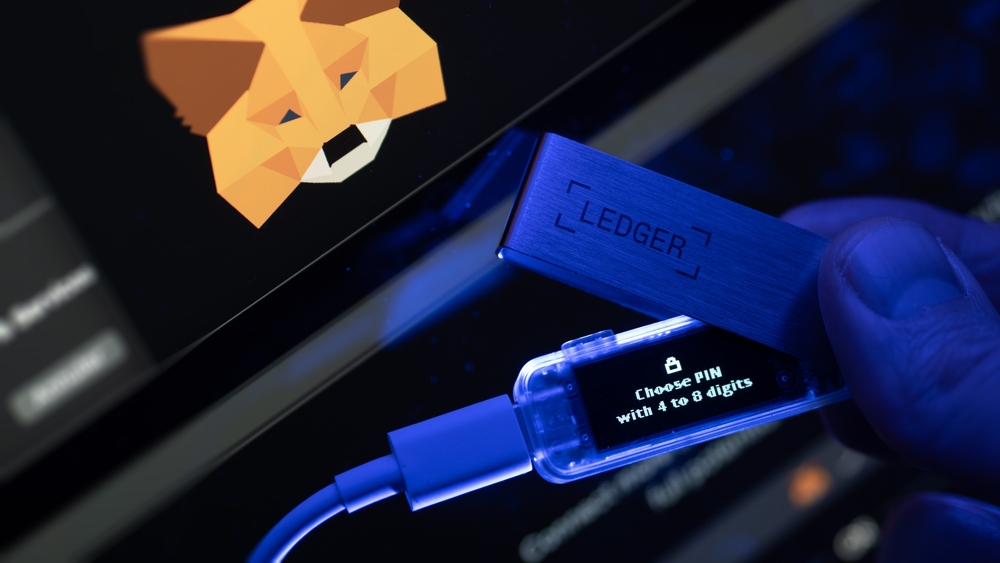 Focus on the screen of an hardware wallet Ledger, the Metamask logo in background. An investor secures her cryptos on a ledger wallet and on Metamask.