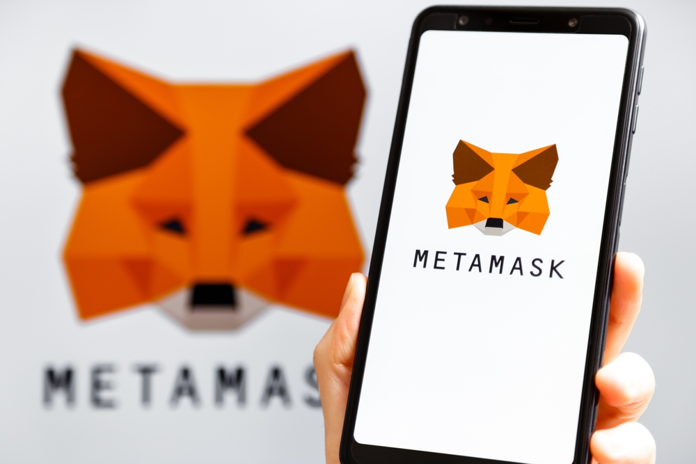 Hand holding mobile with MetaMask app running at smartphone screen with MetaMask logo at background.