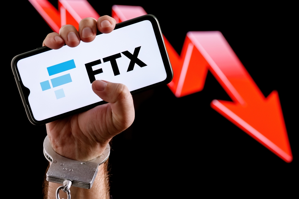 FTX is cryptocurrency exchange. Handcuffed hand squeeze smartphone with FTX logo on screen against background of red down arrow.