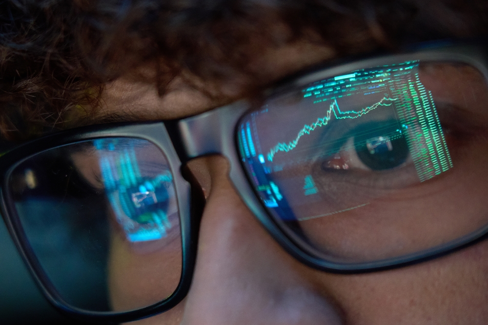 Young indian business man trader wearing glasses looking at computer screen with trading charts