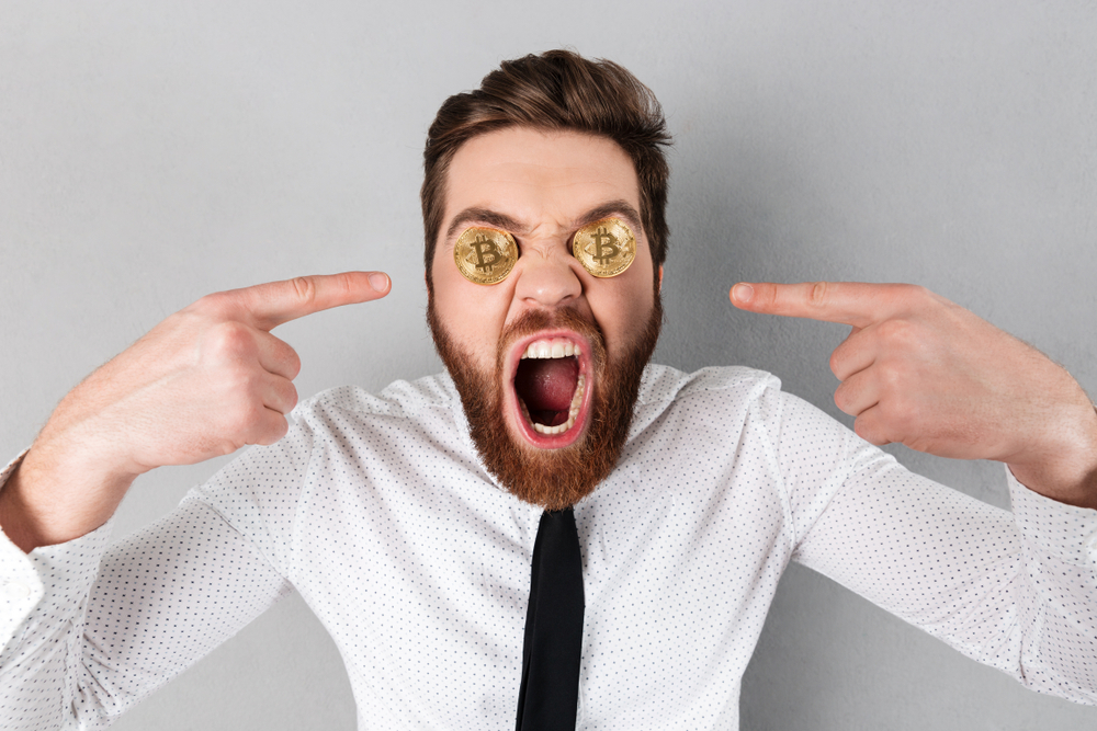 Close up of a crazy businessman with bitcoins in his eyes pointing fingers isolated over gray background