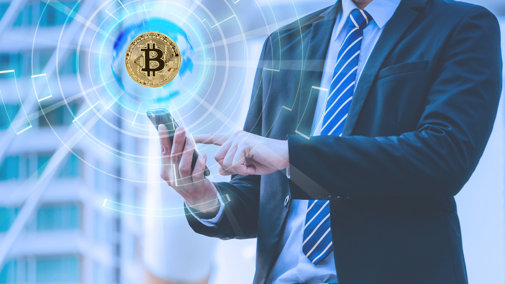 Bitcoin concept with double exposure of Young businessman standing and point to phone and bitcoin global hologram with cityscape background.