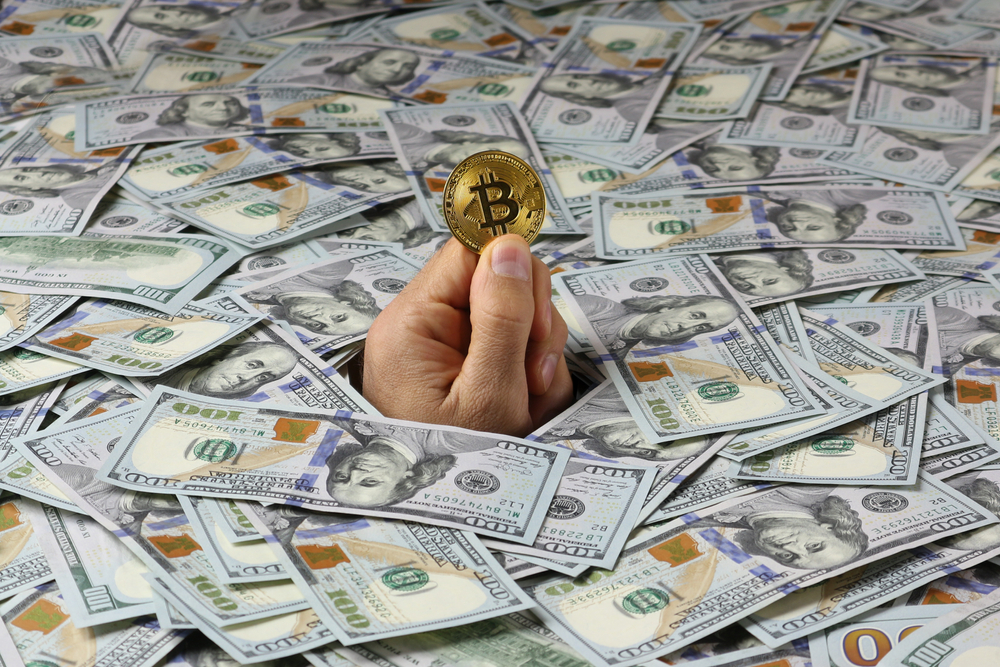 A man’s hand sticks out of a mountain of dollar bills and holds a golden bitcoin. The man drowned in too much money.