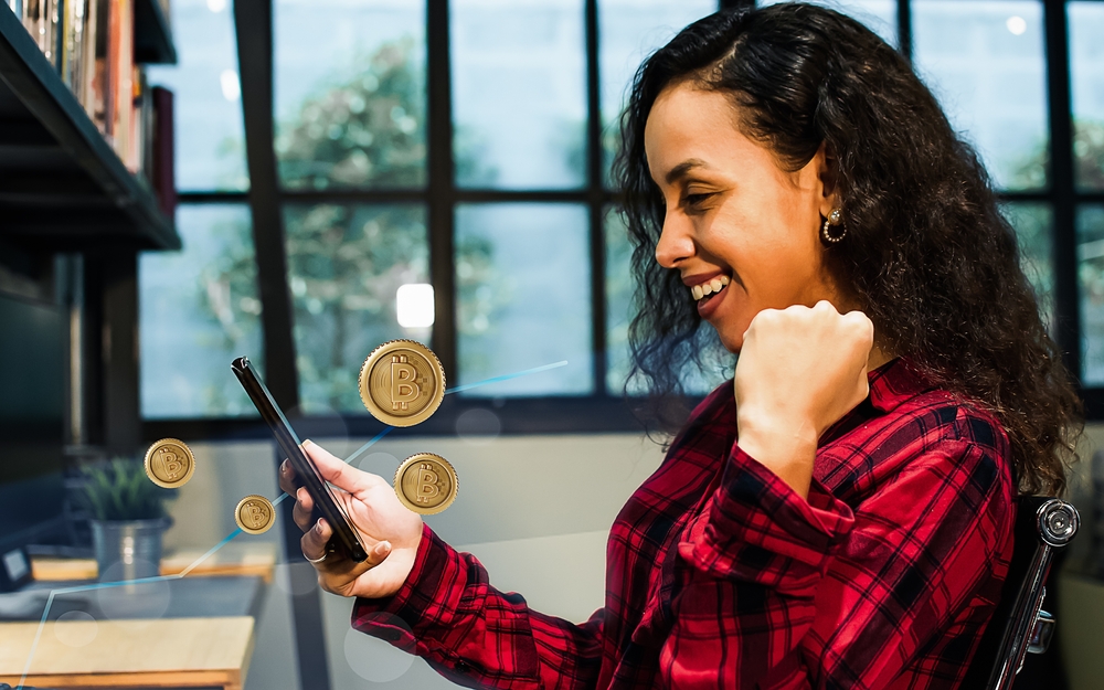 Beautiful cheerful Caucasian woman wearing shirt, holding mobile phone, rasing hand, smiling with happiness and success, invest bitcoin, receiving and win profit. Finance and Investment Concept