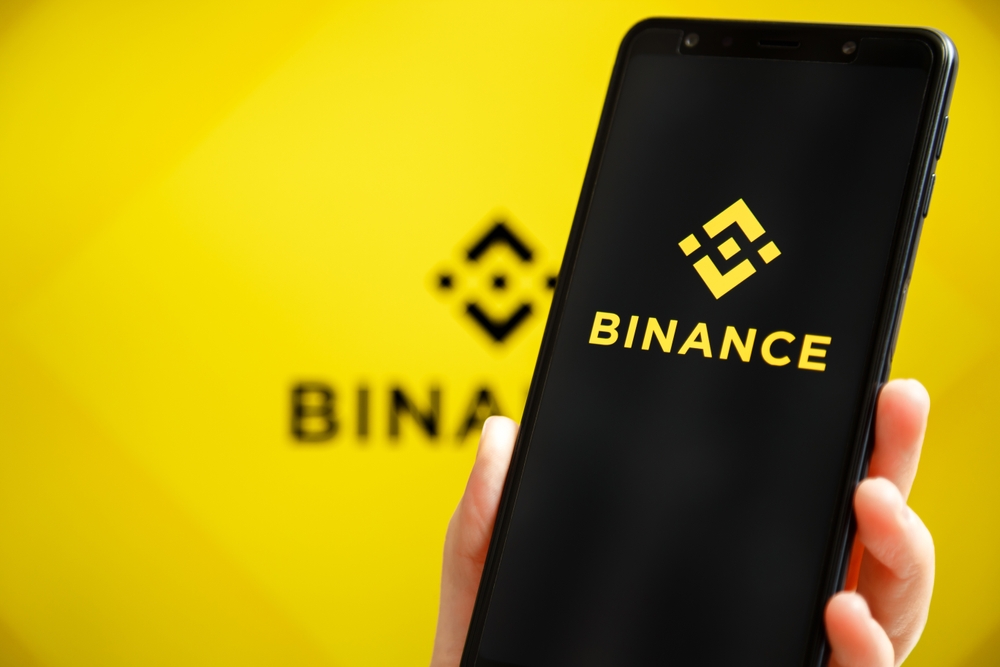 Binance mobile app running at smartphone screen with Binance logo at background.