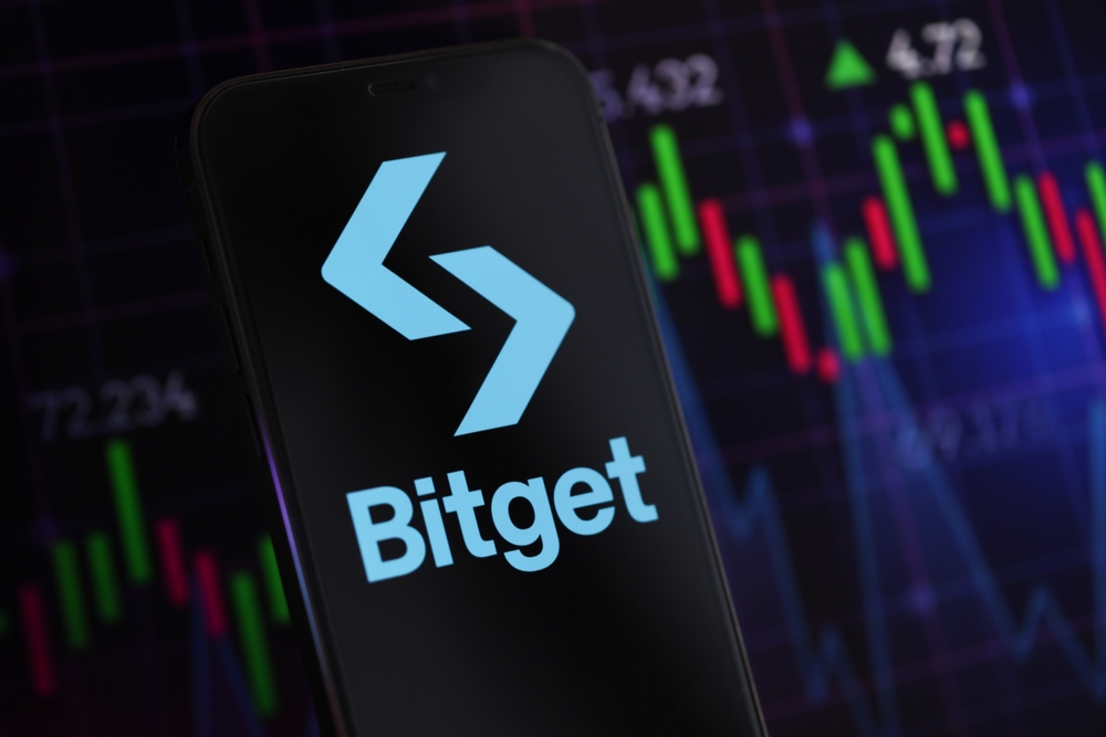 Bitget logo on iPhone display screen and crypto currency value charts. Cryptocurrency exchange portal