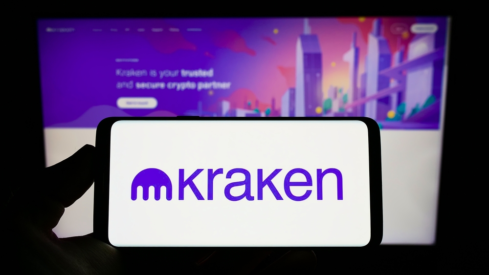 Person holding mobile phone with logo of American crypto company Payward Inc. (Kraken) on screen in front of web page. 