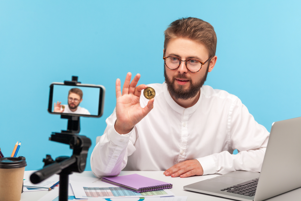 Bearded man blogger holding in hands and showing on smartphone camera golden bitcoin, explaining how to invest in cryptocurrency, mining.