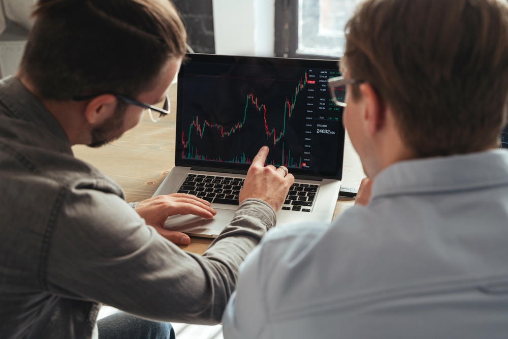 Over shoulder view of trader couch teaching colleague basics of cryptocurrency investments, explaining market trends or consulting his client, analyzing charts of money flow together on pc screen