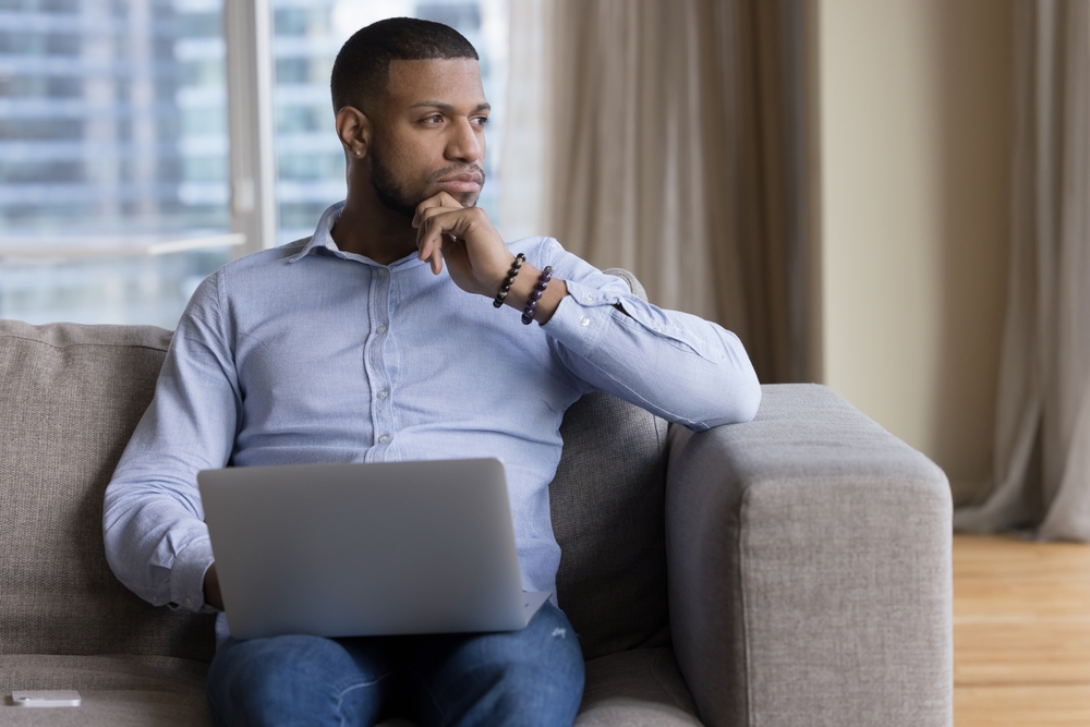 Pensive African guy deep in thoughts, ponders, thinks seated on sofa with laptop staring into distance looks puzzled, uncertain distracted from tech usage, having question or challenge feels undecided