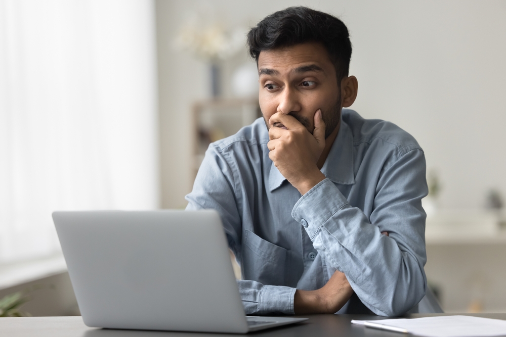Bewildered confused or surprised Indian man sit at desk looking at laptop screen read news