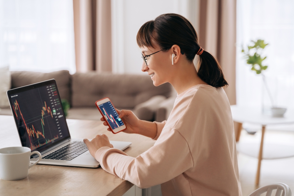 Smiling business woman trader analyst looking at laptop monitor, holding smartphone, wearing earphones.