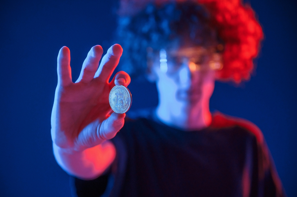 Holding bitcoin. Young man with curly hair is indoors illuminated by neon lighting.
