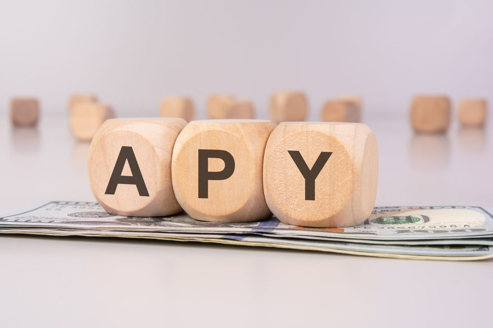 wooden cubes displaying APY rest on a stack of US dollar bills, representing annual percentage yield, interest rates, and financial growth