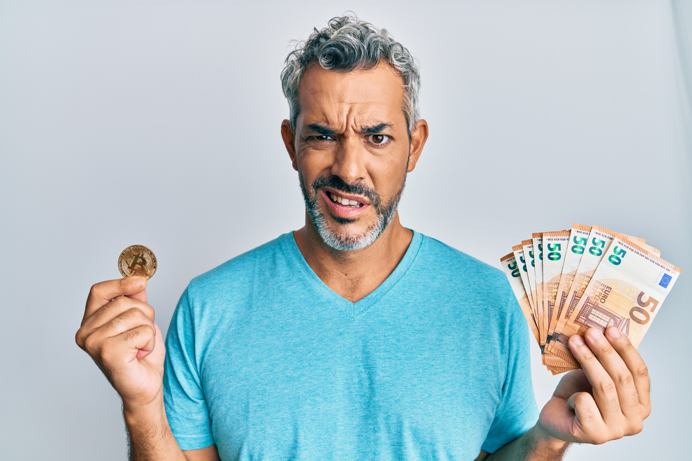 Middle age grey-haired man holding bunch of 50 euro banknotes and bitcoin clueless and confused expression.