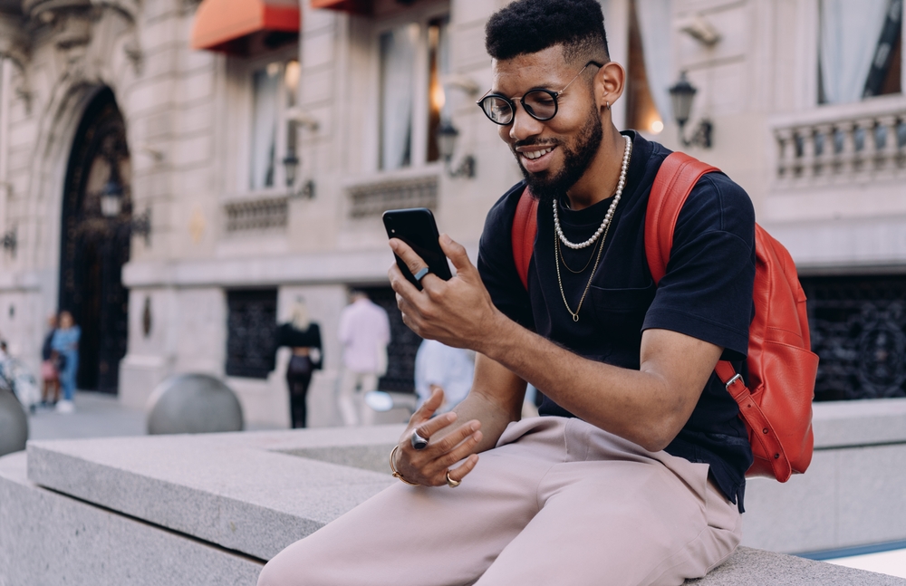 Stylish young Black man holding phone with both hands, sitting against building facade.
