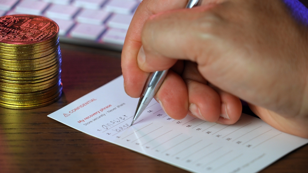 A closeup view of a man writing his secret cryptocurrency wallet recovery "seed" phrase on a note card.